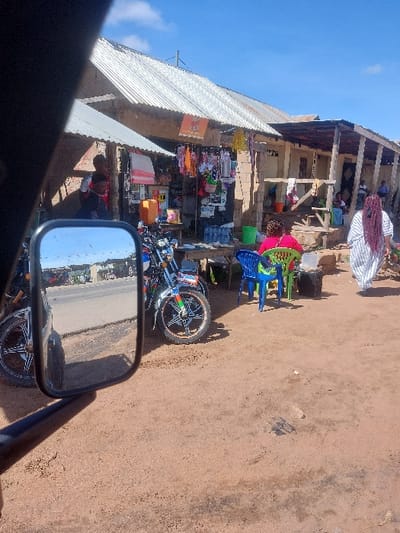 The scene depicts an outdoor commercial area in Kimamba, Tanzania, under bright daylight with a blue sky and scattered white clouds. The photograph is taken from a vehicle, with a black rectangular side mirror occupying the lower left quadrant, reflecting a paved road and distant vehicles.
In the midground, a series of single-story commercial structures with corrugated metal roofs line a reddish-brown dirt pathway. The leftmost visible stall displays various consumer goods, including plastic items and bags, hanging from the storefront. A dark-skinned male, partially obscured in shadow, stands near this stall, wearing a red and white shirt.
Two motorcycles are parked upright on the dirt path. One, blue and white, is on the left. Another, blue with distinctive green and black spoked wheels, is positioned centrally. A wooden table is set between the motorcycles and the path, holding multiple clear plastic bottles of water. Adjacent to the table, a blue plastic chair and a green plastic chair are positioned. A woman wearing a bright pink top is seated on the green chair.
Further to the right, a woman with long, reddish-brown braided hair, dressed in a white garment with dark vertical stripes, walks away from the viewer. Behind her, another building with an open-front porch-like area is visible, where several individuals are seated. The ground throughout the foreground and midground is composed of reddish-brown soil.
