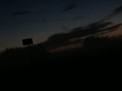 The image depicts a dark, silhouetted landscape at dusk or dawn. A dark, rectangular object, possibly a sign or a panel, is visible on a pole in the left half of the frame. The sky above is a gradient of dark blue and faint orange hues, with some scattered clouds. The foreground and lower portion of the image are entirely black, suggesting a heavily shadowed area or a dense foliage. The overall impression is one of low light conditions, possibly in a rural or semi-rural setting given the silhouette of trees or vegetation. There are no people or discernible actions visible in the image. The location within Воскресенское, Russia cannot be determined from the visual information provided.