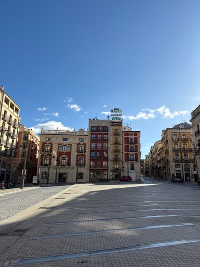 The image depicts a daytime street scene in the city of Alcoy/Alcoi, Spain, under a clear blue sky with minimal scattered white clouds. The foreground consists of a wide, paved plaza area featuring light-colored square cobblestones and parallel metal strips embedded in the surface. Strong sunlight illuminates the scene from the right (east), casting sharp, elongated shadows across the plaza from buildings on the left, oriented roughly towards the left-center of the frame.
Multiple multi-story buildings of traditional European architectural style frame the plaza. On the left, a light-colored building with multiple balconies extends into the background. In the center, a prominent cream-colored building with red decorative paneling around its windows and a white balustrade along its rooftop features dark-framed windows and a ground-floor entrance with glass doors. To its right, another building with a red-tiled facade and dark balconies stands adjacent to a yellow/ochre-colored building. Rooftop signage above these central buildings displays illuminated letters, including "LA SINCÀ" and "CEROL".
Further down a street to the right, buildings with similar architectural styles line the road, which appears to curve into the distance. A small red car is parked on the street in the mid-ground, and a dark grey SUV is visible further down the street. On the left side of the plaza, a smaller cobblestone street or pedestrian area leads off-frame, with a few parked motor scooters visible. No identifiable individuals are present in the immediate foreground or main plaza area. The scene is static, devoid of active interactions.