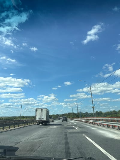 A first-person perspective shot from a vehicle on a highway shows a bright blue sky filled with scattered white cumulus clouds. The highway has multiple lanes and is bordered by a guardrail. Several vehicles are visible on the road, including a white box truck in the foreground and a dark-colored car in the middle distance. Streetlights and power lines run along the side of the highway. A blue sign with white Cyrillic text reading "МОСКВА" (Moscow) is mounted on a pole. The surrounding landscape appears to be rural, with trees and vegetation visible in the distance. The asphalt of the road shows some wear and tear, including cracks and patches. The image is taken on a sunny day.