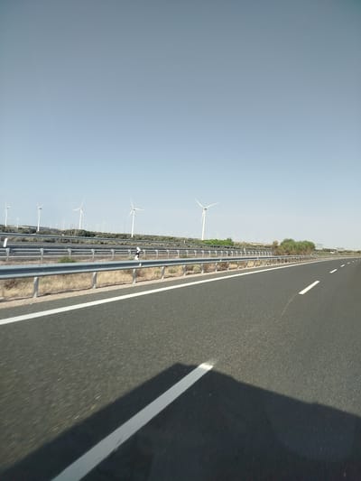 The image depicts a highway landscape with numerous wind turbines visible in the distance. A clear blue sky occupies the upper portion of the frame. In the foreground, the asphalt of the highway is visible with white lane markings. A metal guardrail runs along the side of the road. Sparse vegetation, consisting of dry grass and bushes, is present between the road and the wind turbines. The overall impression is that of a rural or semi-rural area.