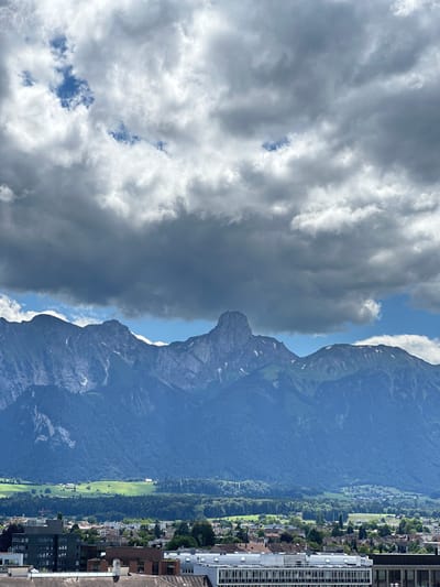 The image captures a panoramic view of the city of Thun, Switzerland, with a dramatic mountain range and cloudy sky as the backdrop. In the foreground, various buildings of different architectural styles are visible, suggesting a dense urban environment. These buildings include a long, modern structure with numerous windows, as well as older structures with pitched roofs. Further back, a lush green landscape with scattered trees and fields stretches towards the imposing mountains. The mountains themselves are rugged and tall, with some patches of snow visible on their peaks. The sky above is filled with a mix of dark, heavy clouds and lighter, brighter clouds, with glimpses of blue sky peeking through. The overall impression is of a city nestled at the foot of majestic mountains under a dynamic sky.