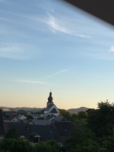 The image shows a panoramic view of the town of Mondercange, Luxembourg, under a clear blue sky with faint wispy clouds. In the center of the image stands a prominent church with a tall, dark steeple topped with a cross. The church is surrounded by a dense collection of residential buildings with dark-colored roofs, characteristic of European towns. Lush green trees are interspersed throughout the landscape, adding a natural element to the urban scene. In the background, rolling hills are visible, suggesting a suburban or semi-rural setting. The overall impression is one of a peaceful, sunlit afternoon in a European village.