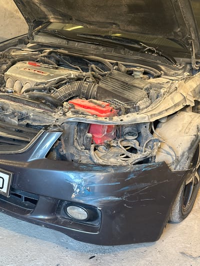 The image shows the front end of a dark-colored sedan, heavily damaged. The hood is open, revealing the engine compartment, which is dusty and cluttered with wires and hoses. The front bumper is scuffed and dented, with visible paint damage. The headlight on the right side is missing, and the surrounding bodywork is deformed. A red battery is visible in the engine bay. The car appears to be stationary in what looks like a garage or workshop, with a concrete floor and a light-colored wall in the background. The location is International, Kyrgyzstan.