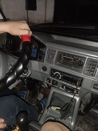 The image captures the interior of a vehicle, likely a car or a small van, from a low, slightly off-center perspective, appearing to be taken by a passenger or someone in the driver's seat looking down and across. The setting appears to be nighttime or a dark indoor environment, indicated by the very dark background visible through the windshield and the reliance on interior lighting to illuminate the cabin.
In the foreground, to the left, a hand is gripping the top of a steering wheel. The steering wheel has a black, somewhat worn cover, with a distinct red section at the top where the hand is holding it. Keys with a keyring are hanging from the ignition, which is located to the right and slightly below the steering wheel.
The dashboard, primarily made of grey plastic, stretches across the width of the frame. On the driver's side, behind the steering wheel, part of the instrument cluster is visible, showing a fuel gauge and a temperature gauge, along with some indicator lights. A distinctive blue and black striped pattern is visible in this area, possibly part of a dashboard mat or design element.
In the center of the dashboard, there are two prominent rectangular air vents, made of grey plastic slats. Below these vents is the climate control panel, which features a horizontal slider for temperature, a rotary dial for fan speed (with markings "OFF", "1", "2", "3"), and mode selection buttons. The word "COOL" is visible on this panel, along with the letters "HL".
Below the climate controls, an aftermarket car stereo head unit is installed. It has a black faceplate, a prominent silver volume knob on the left, and various buttons. Some text, possibly "SL-208" or similar model number, is faintly visible on its display or casing. A coiled black audio jack cable is plugged into the stereo's front auxiliary input and extends downwards. Below this stereo, there is a recessed area or storage slot that appears somewhat worn and damaged.
Further down, between the driver and passenger sides, is a center console unit, also in grey plastic. It features two cup holders and a small open storage compartment. Inside this compartment, a dark green or grey microfiber cloth is placed, along with a crumpled piece of white paper or tissue.
On the floor beneath the dashboard, three pedals are visible: a clutch, a brake, and an accelerator, indicating a manual transmission vehicle. The feet of the person in the driver's seat, wearing blue jeans and dark socks with blue and white accents (resembling athletic socks), are visible near the pedals. To the left of these feet, the black gear stick for a manual transmission is clearly seen. The gear pattern "1 3 5" and "2 4 R" is inscribed on its top. The lower part of another person's legs or knees, wearing light brown trousers, is visible on the far right, suggesting a passenger is also present.
The overall impression is of a somewhat older vehicle interior, showing signs of use and personalization (like the steering wheel cover and aftermarket stereo). The activity appears to be simply sitting in the vehicle, possibly parked, as there's no clear indication of movement, but the hand on the wheel and the keys in the ignition suggest readiness or recent operation.
