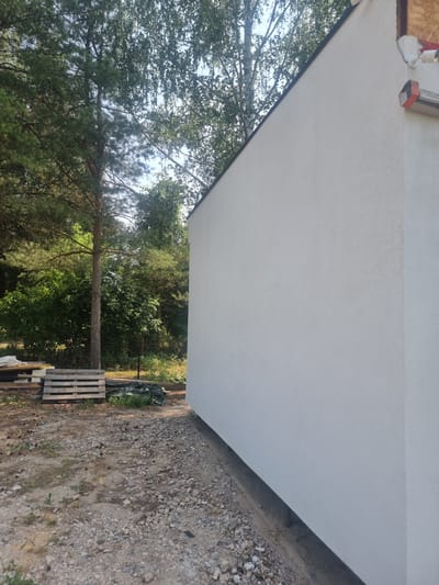 An exterior medium shot shows the side of a white, stucco-finished building with a dark soffit. The building is situated in a wooded area with tall trees and dense green foliage visible behind it. The ground in the foreground is rough and covered with gravel and some scattered pinecones. To the left of the building, wooden pallets and a dark tarp are stacked on the ground. The sky is overcast and partially obscured by tree branches. The scene appears to be in a rural setting, possibly in the city of Szczaki, Poland, indicating an outdoor construction or renovation site.