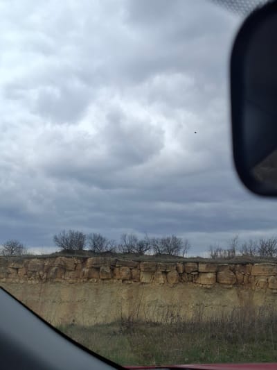 The image presents a natural landscape viewed from inside a vehicle, with parts of the dark dashboard or windshield frame visible in the foreground at the top left and bottom right. The upper portion of the scene is dominated by a cloudy, overcast sky, displaying various shades of grey that suggest diffuse light during daytime. A small, dark, indistinct object, possibly a distant bird or airborne debris, can be seen against the clouds in the upper left quadrant.

Horizontally across the mid-ground stretches a prominent geological feature: a layered rock formation or cliff face. The rocks are light brown and tan, exhibiting rugged textures and distinct, somewhat blocky strata. Along the top of this rock formation and extending into the background are numerous bare deciduous trees, indicative of a late winter or early spring season. The ground and areas atop the cliff are covered with dry, brownish grasses and sparse brush, with some patches of greener vegetation visible.

The overall impression is a somewhat stark and muted rural landscape, consistent with Yablonovo, Russia, under an overcast sky. No people are visible, and no text is discernible. The activity depicted is simply the observation of scenery, likely from a passing vehicle.