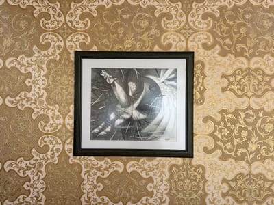 A framed, black and white drawing is displayed on a wall with ornate, gold and cream patterned wallpaper. The drawing, which appears to be a sketch or etching, features a dynamic, abstract composition. The foreground of the drawing shows a horse-like head with an open mouth, surrounded by sharp, intersecting lines that suggest a chaotic or energetic scene. Further into the drawing, there are hints of draped fabric or flowing forms with distinct patterns, possibly representing a dress or other decorative elements. The drawing is signed in the bottom right corner with what appears to be an Arabic inscription. The frame is dark wood. The wallpaper has a repeating pattern of floral and scrolling motifs. The lighting is bright and even, casting no significant shadows. The overall impression is one of an art display in an interior setting.