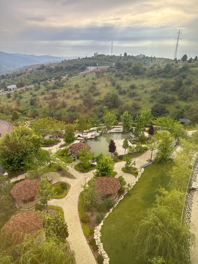 The image depicts an aerial view of a park-like setting with a small pond, several hexagonal gazebos with brown shingled roofs, paved walkways, and lush greenery. The park appears to be situated on a hillside overlooking a valley with rolling hills in the distance, some of which are covered in trees and vegetation. Power lines and pylons are visible on the hillsides, suggesting an urban or suburban periphery. A road is visible in the upper left quadrant of the image, leading into the park. The weather appears overcast, with a soft, diffused light. No people are clearly discernible in the image. The scene suggests a recreational area designed with natural elements and man-made structures for leisure.