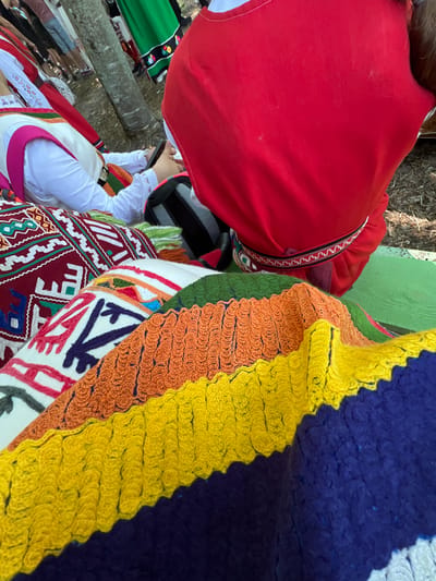 This image captures an outdoor scene in Bolyarovo, Bulgaria, centered on traditional textiles and individuals in customary attire. In the foreground, a stack of vibrant, hand-knitted or crocheted blankets is displayed, featuring distinct horizontal stripes in dark blue, yellow, orange, and green, each stripe exhibiting a textured, wavy pattern. Beneath these are other woven textiles, likely cushions or rugs, adorned with complex geometric patterns in white, red, green, and dark blue on maroon and white bases.
In the mid-ground, two individuals are prominent. On the left, a person is seen from the back, wearing a white long-sleeved shirt with embroidered cuffs, a white apron-like overgarment, and a colorful striped sash (pink, green, white). This individual holds and observes a dark-colored smartphone. To the right, a person is depicted from the upper back, wearing a bright red garment, possibly a vest or top, over a white layer, cinched by a narrow belt featuring white, black, and red geometric patterns. This person is positioned on a green painted wooden surface, possibly a bench or table.
The background contains several blurred figures, some standing, suggesting a gathering. Visible attire includes a green skirt with a decorative black band on a figure in the background right, and a red top with white embroidery on a figure in the far left background. A tree trunk is visible in the mid-ground, indicating a natural, outdoor setting with visible soil and scattered ground cover.