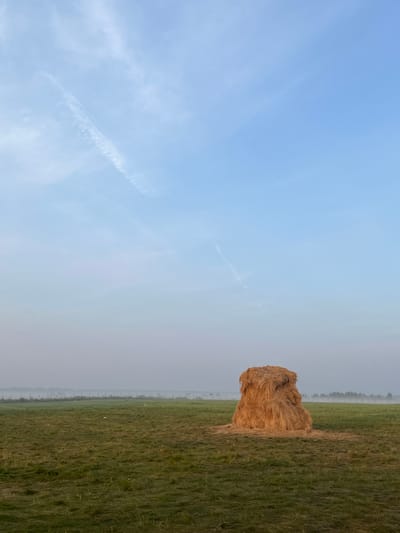 The image depicts a rural landscape in the city of Surakovo, Russia, under a pale blue sky with thin wispy clouds. In the foreground, a lone, large haystack stands in a vast, green grassy field. The grass appears recently cut in some areas, with patches of stubble. In the background, a faint mist or fog hovers over the horizon, obscuring a line of trees and a fence. The overall atmosphere is calm and serene, suggestive of early morning or late evening light. There are no people or animals visible in the scene.