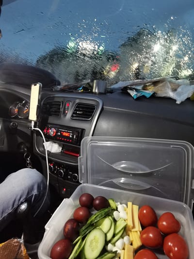 The image depicts the interior of a car, possibly stationary, due to heavy rain obscuring the windshield and streetlights. A person wearing jeans is partially visible in the driver's seat. On the dashboard, there is a mobile phone mounted on a holder, connected with a white cable to a car stereo displaying "106.2". A metal mug, snack wrappers, and other miscellaneous items are scattered on the dashboard. A clear plastic food container with a lid resting on top is in the foreground, containing food items, including tomatoes, cheese cubes, cucumbers, and dark red plums. Given the city is Baskan, Russia, the environment outside the car is likely urban, though visibility is heavily impacted by rain.