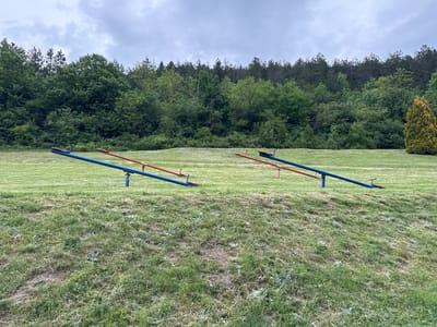 Two blue and red seesaws stand on a grassy field in Despotovac Municipality, Serbia. A line of trees borders the field. The sky is overcast.
