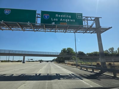 The image shows a view from a vehicle driving on a highway. Above the highway, a green overhead sign indicates "INTERSTATE 80" on the left and "INTERSTATE 5 Redding Los Angeles EXIT 86" on the right. A yellow sign below indicates "ONLY". The sky is blue and clear with some sun glare. In the distance, there are trees, other vehicles, and a pedestrian overpass. The highway appears to be in a suburban or rural area.