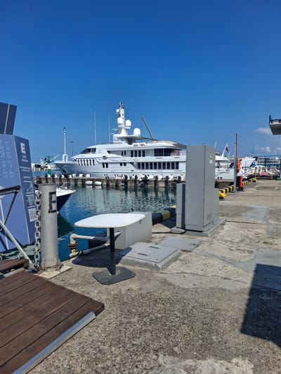 The image depicts a marina scene under a clear blue sky. In the background, a large white yacht is docked, its multiple decks and superstructure visible. Adjacent to the yacht, other smaller boats are moored. The foreground shows a concrete pier with various elements. A grey metal utility box stands prominently on the pier, with yellow and black striped bollards along the edge. A round white table with a black base is also present on the pier. To the left, a wooden walkway or ramp leads into the scene, adjacent to a blue informational sign with text and coordinates, including "43° 34' 50.9°N" and "39° 43' 07.0°E". The water in the marina is calm and reflects the sky and surrounding structures. The overall environment suggests a sunny day at a harbor. The coordinates provided on the sign correspond to a location in Sochi, Russia, not Guzeripl', Russia.