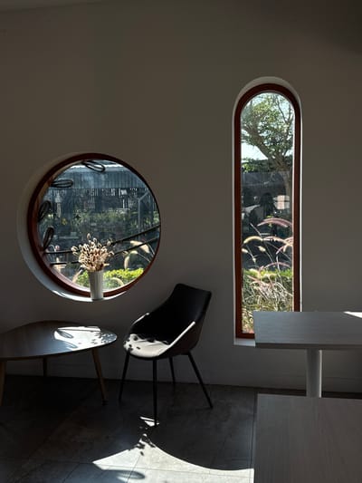 An interior shot from San Phra Net, Thailand, depicts a minimalist room bathed in natural sunlight. The space features light-colored walls and a tiled floor with visible grout lines. On the left, a circular window with a prominent dark wooden frame and white outer trim looks out onto an overgrown exterior garden area with various green plants and what appears to be a metal fence or railing in the mid-ground. A white fluted vase containing dried, light-colored seed heads or botanicals rests on the sill of this window. Adjacent to the circular window, a small, round wooden table stands on two slender legs, partially illuminated by sunlight casting leaf shadows. A modern-style chair with a black upholstered seat and back and four thin black legs is positioned next to the table, also receiving sunlight and casting shadows. On the right, a tall, narrow arched window with a dark wooden frame provides a view of lush green foliage, a tree trunk, and vibrant feathery grass-like plants (Pennisetum) outside, brightly lit by the sun. In the foreground on the right, a rectangular light-colored table with a white pedestal base is visible, and a portion of another similar table is seen in the bottom right corner. No individuals are present in the scene.