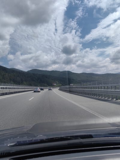 The image shows a view from inside a vehicle driving on a multi-lane highway, likely in a mountainous region, given the dense forest covering the hills in the background. The highway has clear lane markings and is bordered by a metal guardrail. Several cars are visible on the road ahead, traveling in the same direction. The sky is partly cloudy with patches of blue sky and white clouds, suggesting daytime. The foreground of the image is dominated by the dashboard and windshield of the vehicle, with a slightly blurred view of the road and scenery beyond. There are no identifiable people or specific landmarks that would pinpoint the location to Potop, Bulgaria, or any other specific city.