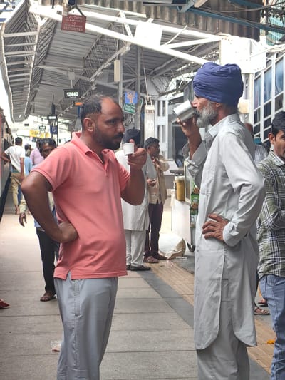 The image depicts two men on a train platform in Khandwa, India. The man on the left, with a beard, is wearing a pink polo shirt and grey trousers, holding a small white cup and gesturing with his hand. The man on the right, wearing a blue turban and a light-colored kurta, is drinking from a metal cup. The platform is covered by a metal roof supported by white pillars, with overhead signage and tracks visible. Other individuals are present in the background, with luggage and vendors on the platform. The scene appears to be an active railway station.