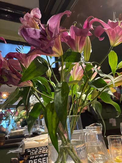 The image shows a bouquet of pink lilies in a transparent vase filled with water. The flowers are predominantly pink with some white and yellow accents. Green leaves surround the stems. Behind the vase is a collection of empty glass containers and a stack of white napkins with black lettering. The reflection in a mirrored surface shows more flowers and a partial view of what seems to be a restaurant interior. Due to the limited information and the absence of clear landmarks, a specific location within Aberystwyth, United Kingdom cannot be determined. The scene is static, depicting an arrangement rather than an action.