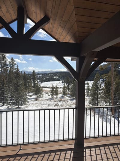 A view from a wooden structure with supporting beams, likely a porch or balcony, overlooks a snowy landscape. The scene features a forest with bare trees interspersed with evergreens and a snow-covered area resembling a frozen lake or field. A dark railing runs along the right side. The sky is visible with scattered clouds. The wooden structure's architecture includes triangular bracing.