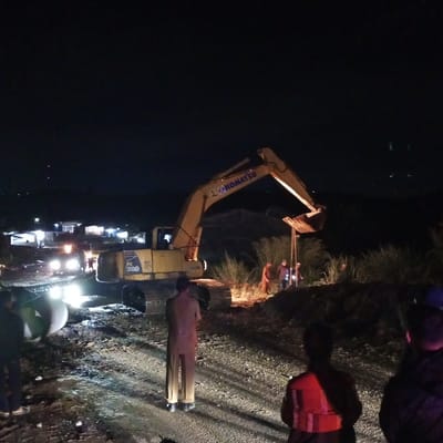 A night scene in Bintangpermata, Indonesia depicts a construction site with an excavator and several individuals. The excavator, a yellow Komatsu PC 200, is positioned centrally. Several people are visible, some wearing high-visibility vests. The ground appears to be dirt and gravel, wet and reflective due to lighting. In the background, there are structures with illuminated windows, suggesting a settlement or residential area. The overall lighting is dim, with artificial lights from the construction equipment and surrounding buildings illuminating the scene. The sky is dark, indicating nighttime.