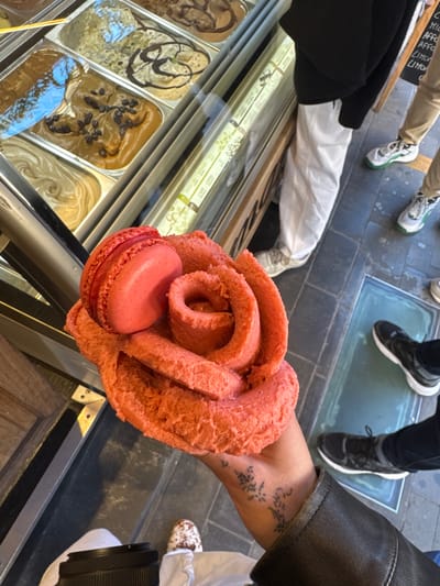 A person holds a pink rose-shaped gelato with a matching macaron in front of a display case containing various flavors of gelato. The gelato display is likely in a shop in Sorrento, Italy, as rose-shaped gelato is a common offering there. The person holding the gelato wears a dark jacket and has a tattoo on their wrist. In the background, there are glimpses of other people's legs and shoes, indicating that they are likely customers in the shop. The floor is tiled and has glass panels.