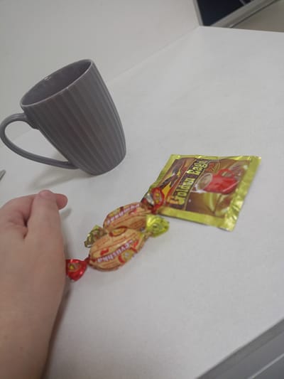 A close-up shot shows a person's hand on the left side of the frame, reaching towards two individually wrapped candies. The candies are golden with red accents, and appear to be twisted at the ends. To the right of the candies is a small, golden packet with the text "Golden Eagle 3-in-1 Classic" visible, along with an image of a red mug of coffee. Behind the candies and packet, a ribbed, grey mug is positioned with its handle facing left. The items are resting on a white, textured surface, likely a desk or table. The lighting suggests an indoor setting, possibly during the daytime. The background is out of focus, with a hint of a grey and white surface and a darker object in the upper right corner, possibly a shelf or another piece of furniture. The scene appears to be a simple display of a beverage packet and some sweets.
