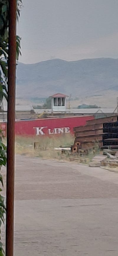 The image depicts a view of a large red structure with "K LINE" painted in white letters on its side. A small, white, elevated structure with a red roof and windows is visible on top of the red structure. In the foreground, a dark wooden post with green leaves is partially visible on the left. The background shows a barren landscape with distant hills under an overcast sky. The scene appears to be an industrial or port area. The location is identified as Ohangaron, Uzbekistan.
