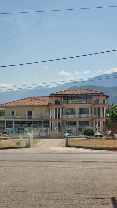 A three-story building with an orange-tiled roof is pictured in Kalambaka, Greece. The building's facade is a mixture of beige and peach colors, with large windows and balconies on the upper floors. The ground floor appears to be a commercial space with storefronts displaying products and signage, including "Berling" and "ΣΠΑΝΟΣ". A metal gate and fence are visible in front of the building, with a paved driveway leading into the property. Several vehicles, including a white van and a white SUV, are parked on the property. The background shows a range of mountains under a clear blue sky with a few scattered clouds. Overhead power lines are visible in the foreground.