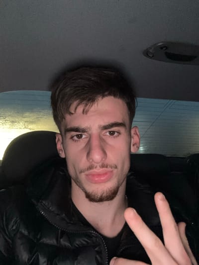 A young man is captured in a close-up, self-portrait style photograph. He appears to be in his late teens or early twenties, looking directly into the camera with a neutral to slightly serious expression. He has short dark hair with a fringe and a neatly trimmed dark beard and mustache, styled in a chinstrap fashion. His eyes are dark brown.
He is wearing a black, possibly shiny, puffer jacket, zipped up, with a subtle silver-colored chain visible around his neck. His right hand is raised into the frame, forming a "peace sign" or "V-sign" gesture with his extended index and middle fingers.
The background is dimly lit and predominantly dark, suggesting an indoor or nighttime setting. To his left, a dark, featureless surface is visible. In the upper left portion of the background, a dark, elongated object, possibly a handle or part of an interior fixture, can be vaguely discerned. To his right, a lighter area with a faint linear or textured pattern is present, possibly a window reflecting light or a textured wall. The environment suggests an enclosed space, such as a vehicle or a room with subdued lighting. There is no visible text in the image.