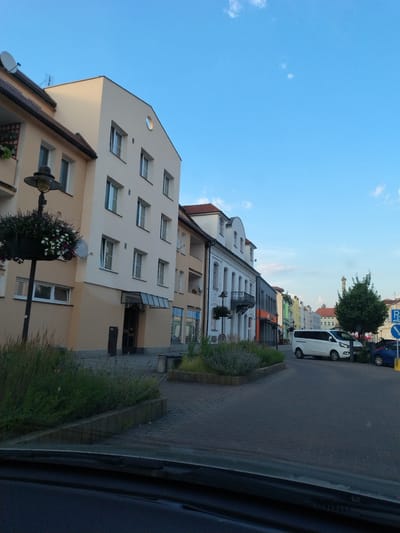 The image shows a street scene in Strakonice, Czechia. On the left, a light yellow building with white trim and several windows is visible. A lamppost with a hanging flower basket stands in front of it. To the right, a row of buildings of varying heights and colors line the street. The buildings are a mix of residential and commercial. A white van is parked on the street, and a blue car is parked behind it. A blue and white parking sign is visible on the right side of the image. The sky is clear and blue with a few scattered clouds. The street is paved with cobblestones. The scene appears to be from a town square or a main street.