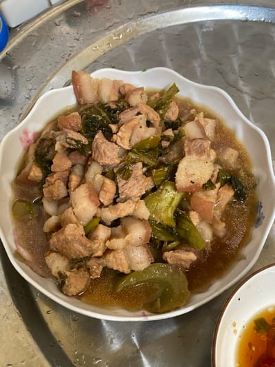 The scene depicts a cooked meal presented in a white, ceramic bowl with a scalloped edge, located in Đak Đoa, Vietnam. The bowl rests on a wet, reflective metallic surface, consistent with a stainless steel kitchen sink or countertop, evidenced by numerous water droplets. The dish consists of diced pork belly, identifiable by distinct layers of lean pinkish-brown meat and off-white fat, mixed with green leafy vegetables, likely mustard greens, all immersed in a light brown, translucent broth. A small, partially visible white bowl containing a reddish-orange dipping sauce with green flecks is situated in the lower right portion of the frame. A blue object is partially visible in the upper left background. No individuals or active interactions are present.