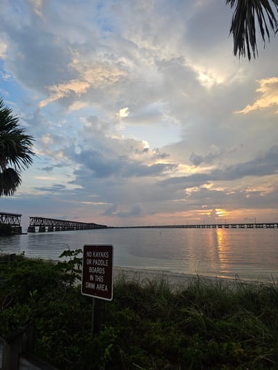 The image is a landscape view of a beach scene in Big Pine Key, United States, taken during sunset. The sky is filled with clouds illuminated by the setting sun, casting a golden hue on the water. A bridge stretches across the water in the distance. In the foreground, there is vegetation and a sign that reads "NO KAYAKS OR PADDLE BOARDS IN THIS SWIM AREA." No people are visible.