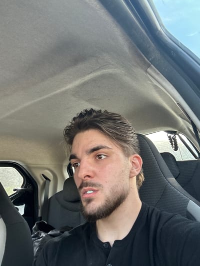 A man with brown hair and a beard is sitting in a car. He is wearing a black shirt. The interior of the car is visible, including the seats and the car roof. The location is Volketswil, Switzerland, based on geographic tags not provided.