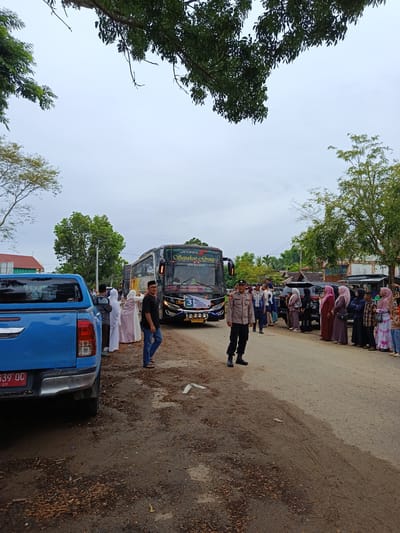 A group of people are gathered on a dirt road in Meureudu, Indonesia, near a large bus. The bus is black with colorful decals and reads "JETBUS" and "Sepsaka" above the windshield. A blue pickup truck is parked on the left side of the road, its rear visible. Several people are standing in a line on the right side of the road, some wearing traditional Islamic clothing. A police officer in uniform is standing near the bus, looking towards the camera. The sky is overcast, suggesting a cloudy or possibly rainy day. The trees are lush and green, indicating a tropical or subtropical climate. The ground is a mix of dirt, gravel, and scattered dry leaves. The scene appears to be a pickup or drop-off point for the bus, possibly a group traveling together. The presence of the police officer suggests an organized event or a need for security. The mood appears calm and orderly. There is text on the license plate of the truck, "B 939 QC", and on the bus, including "JETBUS", "Sepsaka", and a number "3". There is also text on a banner on the front of the bus, though it is difficult to read.