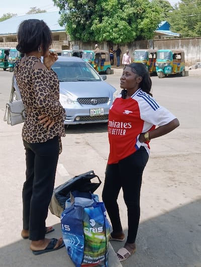 This image captures an outdoor urban or semi-urban scene in Communauté rurale de Payar, Senegal, under bright daylight.
**People:**
Two adult females are in the foreground, facing each other. The female on the left is dark-skinned, viewed from her back-left. She wears a brown and black leopard-print long-sleeved top with cut-out shoulders, black trousers, and dark flat sandals. A light grey quilted handbag hangs from her left shoulder, and her right hand is positioned near her mouth. The female on the right is dark-skinned, viewed from her front-left, looking towards the first female. She wears a red and white Adidas Arsenal football jersey with "Emirates FLY BETTER" sponsorship, black leggings, and light beige slide sandals. A gold-colored watch is on her left wrist, and her hands are on her hips. In the background, several indistinct male figures are standing or walking near vehicles and buildings.
**Objects:**
On the paved ground between the two females are a black open tote bag and a larger blue shopping bag bearing "ORIS" branding and a landscape graphic. A silver Toyota Corolla sedan, with a Nigerian license plate "DR-131-AA", is parked on an asphalt road behind the females. Several three-wheeled motorized rickshaws (tuk-tuks) in green, yellow, and blue colors are parked further back; one displays "PTN 6333".
**Environment and Actions:**
The two females are standing on a concrete surface, engaged in an apparent conversation or interaction. Behind them, an asphalt road leads to a background featuring a concrete wall, green trees, and light-colored buildings with blue roof accents. The sky is clear, and the scene is brightly illuminated, casting visible shadows.