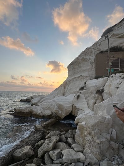 The image depicts a scenic view of the Rosh HaNikra grottoes, located in Mate Asher Regional Council, Israel. A person's hands are visible in the lower right corner, holding a smartphone, possibly taking a photograph or recording a video of the scene. The foreground is dominated by a collection of large, weathered rocks and boulders scattered along the shoreline. The sea, with its gentle waves, meets the rocky coast on the left side of the image. Behind the rocks, a dramatic white cliff face rises sharply. The cliff exhibits distinct horizontal striations, indicative of its geological formation. A small, stone-clad building with a metal gate and some signage is integrated into the cliff face, slightly above the waterline. The sky above is partly cloudy, with soft, warm hues of orange and pink from a sunset or sunrise. Sparse vegetation grows on the slopes of the cliff.