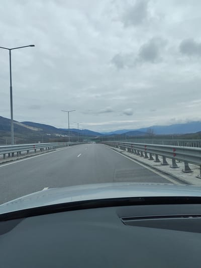 The image depicts a view from inside a car driving along a multi-lane highway in Novachene, Bulgaria. The highway is elevated, possibly a bridge or overpass, with guardrails on both sides. Streetlights line the road. In the background, there are rolling hills or mountains under an overcast sky. The car's dashboard and windshield are visible in the foreground. There are no people visible, only the road and the surrounding landscape.