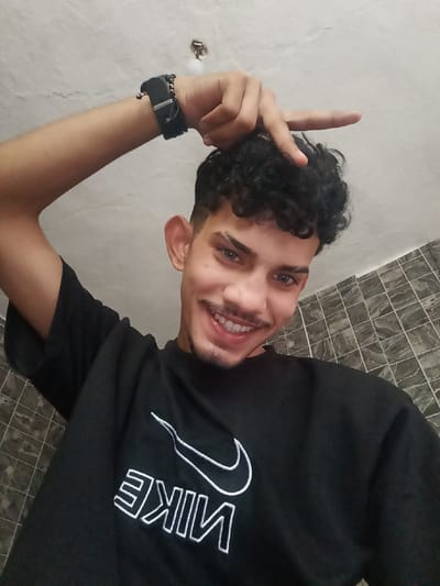 A young man with curly dark hair and light blue eyes smiles at the camera, holding his right hand up to his forehead with his index finger extended. He wears a black t-shirt with a white Nike logo. The background consists of a tiled wall and a white ceiling with a light fixture. The image appears to be a selfie taken indoors.