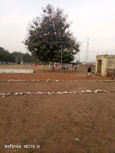 The image depicts an expansive outdoor scene in Ayere, Nigeria, characterized by a dry, dusty landscape. In the foreground, reddish-brown earth is prominent, sparsely covered with dry grass, and marked by two parallel lines of white-painted rocks, possibly indicating a pathway or boundary.

A large, mature tree with dense foliage stands centrally in the mid-ground. Beneath its shade, a group of people is gathered, with some individuals appearing to be seated under a rudimentary shelter. Beyond the tree, a wide, open field serves as a sports ground. Here, numerous people, many distinguishable by their red, white, or blue jerseys, are engaged in what appears to be a soccer match, with goalposts visible on the field.

To the right, a weathered, light-colored building, possibly constructed from concrete or stucco, features a wooden barrier or gate near its entrance. A man, dressed in a dark top and light shorts, is seen walking from right to left in the mid-ground, passing this building while holding a small, dark, round object in his hand.

The background reveals a tall communication tower on the right and indistinct distant hills or structures, all under a bright, overcast sky that suggests either early morning or late afternoon with a hint of haze. The image bears a "Infinix NOTE 8i" watermark in the bottom left corner. The scene portrays a community engaging in recreational activities within a rural or semi-urban environment.