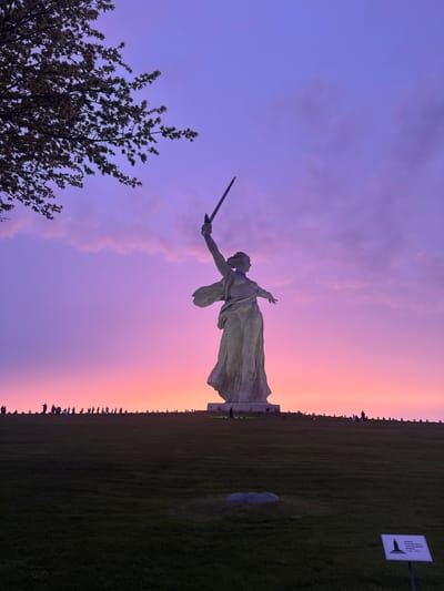 The image shows the Motherland Calls statue in Volgograd, Russia, against a vibrant sky. The sky has a gradient from pink to purple. In the foreground is a tree. The green grass field stretches from the statue toward the right of the image, lined with people. A sign is visible on the right side.