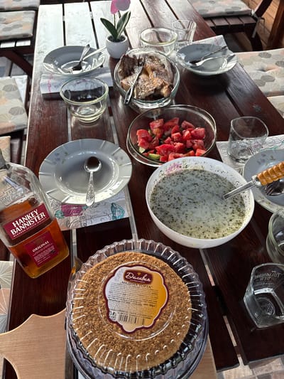 The image depicts an outdoor dining table set with various food and beverage items in Polena, Bulgaria. The central object is a rectangular wooden table composed of dark brown planks, with lighter, weathered planks at the far end. Sunlight illuminates the table, creating reflections.
On the table, from left to right:
A clear glass bottle of "Hankey Bannister Original Blended Scotch Whisky" is positioned at the bottom left, partially obscuring a patterned cushion on a chair.
Next to it, a round, packaged cake, labeled "Деливио МЕДЕНА ТОРТА" (Delivio Honey Cake) in Cyrillic, with a crumb topping, sits in a clear plastic container. The label indicates a weight of "НЕТО: 350g" and a price of "4.80€".
A large white bowl containing a light green, creamy soup or dip with dark green herb speckles is prominently displayed, with a serving spoon resting inside.
To the right of the soup bowl, a clear glass tumbler with "Tullamore D.E.W. Irish Whiskey" embossed on its base is present.
Further up, a clear glass bowl holds chopped red tomatoes and green cucumbers.
Above the salad bowl, a white plate with a floral pattern holds a serving fork and spoon.
At the top center, a clear glass bowl contains several pieces of cooked fish or meat, with a serving fork inserted.
To the left of the fish bowl, an empty clear glass tumbler and a smaller empty clear glass bowl are visible.
Another white plate with a floral pattern, holding two spoons, rests on a patterned paper napkin.
At the top right, a small white potted plant with a single pink artificial flower adds a decorative element.
Several empty patterned plates and additional empty clear glass tumblers are scattered across the table. Patterned cushions on unseen chairs are visible along the left and top edges of the frame, suggesting a gathering. No people are visible in the scene.