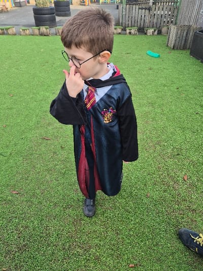 A young male child, approximately 4-6 years old, is depicted standing outdoors on artificial green turf. He has short brown hair and is wearing round, black-rimmed glasses. He is dressed in a dark-colored costume resembling a Hogwarts student uniform, featuring a dark robe with a maroon lining, a white shirt, and a striped tie (maroon and gold/yellow). A crest, likely Gryffindor, is visible on the left side of his robe. His right index finger is positioned at or near his nose. He wears dark shoes.
In the background, to the left, three dark-colored tires are stacked vertically. Behind these are multiple wooden stumps or logs arranged on the ground. A wooden slatted fence extends across the mid-ground. Further right, a dark green, elongated object lies on the turf. The tip of a black shoe with yellow laces is visible in the bottom right corner of the frame. The scene appears to be an outdoor play area, likely within a school or nursery setting in the city of Hereford, United Kingdom, though a precise address is not discernible from the image content.