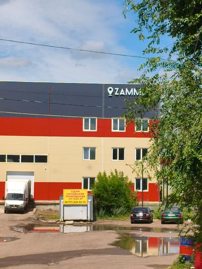 This image depicts an exterior view of a modern industrial building in the city of Zaşağan, Kazakhstan. The building features a distinctive color scheme with a dark gray upper section, a bright red middle band, and a cream-colored lower section. Multiple windows are visible across the building's facade. A large white truck is parked near a loading bay on the left side of the image, adjacent to a small utility structure with a yellow sign. This sign contains text in Cyrillic, indicating "RENT WAREHOUSE SPACE FROM 500 M²" and a phone number. Two cars, a dark sedan and a green sedan, are parked in the foreground on the right. The ground in front of the building and the parking area is paved and shows several large puddles, suggesting recent rainfall. Lush green foliage, including a large tree in the foreground, frames the right side of the scene, and the sky above is partly cloudy. The overall impression is one of an active commercial or industrial facility.