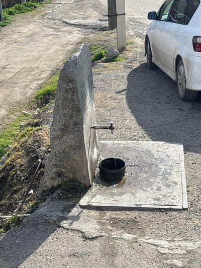 The scene, located in Lehvaz, Armenia, depicts an outdoor water fountain on the side of a road under direct sunlight. A large, irregularly shaped, light gray stone slab stands vertically, acting as the primary structure of the fountain. From the lower-mid section of this stone, a metal faucet with a cross-shaped handle protrudes horizontally, from which a steady stream of water flows downward. This water is collected in a dark, cylindrical pot or bucket positioned on a wet, light gray concrete slab directly below the faucet. The surrounding ground, a mix of asphalt and concrete, shows evidence of moisture. To the immediate left of the stone fountain, a raised dirt and gravel shoulder features patches of green grass and a segment of white plastic tubing. To the right of the fountain, the rear section of a white vehicle, possibly a sedan or small station wagon, is parked on a gravel or asphalt surface. Further right and slightly behind the fountain, a light-colored utility pole is visible. Distinct shadows are cast across the ground, including a large, indistinct shadow on the road surface to the right of the fountain and car. The background consists of an unpaved or gravel path. No individuals are present in the image.