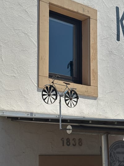 The image captures a portion of a building's facade in Sankt Wendel, Germany. A white, textured wall forms the primary background. A window with a beige stone frame is centrally located, reflecting a blue sky and some overhead structures. Below the window, a decorative metal bicycle ornament is mounted on a vertical rod. The bicycle has two wheels with spokes and a basket. Beneath the bicycle, a metal awning or gutter system is visible, attached to the building with screws. Further down, the numbers "1838" are embossed on the wall, suggesting a historical context or address. To the right, a circular sign with text that appears to be "CUSTOM PAINTING & TATTOO" is partially visible. The overall lighting suggests a bright, sunny day.