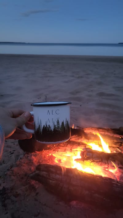A person's hand holds a white enamel mug with a dark rim and handle. The mug features a design of dark green pine trees against a white background, with two lines of text above the trees that appear to be in Cyrillic script. The mug is positioned in front of a crackling campfire on a sandy beach. The fire is large and bright, with visible flames and glowing embers. In the background, a calm body of water stretches to the horizon, meeting a pale blue sky that suggests either dawn or dusk. The overall scene depicts a cozy and tranquil moment by the water, with the campfire providing warmth and light. The location is the city of Zaostrov'e, Russia.