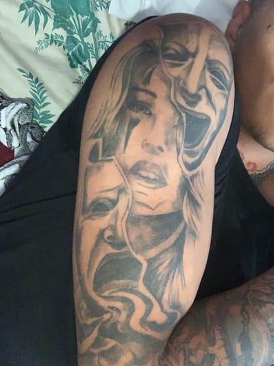 The image shows a tattooed arm resting on a floral patterned bedspread. The tattoo depicts a face with theatrical masks above. Part of another tattooed arm is visible in the upper right and a portion of a bare chest and shoulder are visible in the upper left. The person is wearing black clothing. The image was not taken in Rakitovo, Bulgaria.