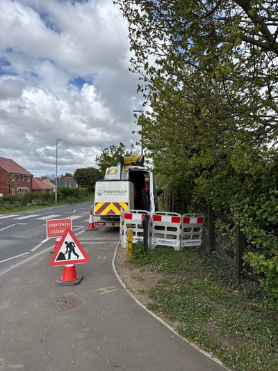 The image depicts a roadside scene in Mid Suffolk, United Kingdom. A white van, labeled "FIBRESOF SOLUTIONS", is parked partially on the paved sidewalk and the adjacent road. The rear doors of the van are open, revealing equipment inside. Two red and white barrier sections, also labeled "FIBRESOF", are positioned behind the van. A traffic cone with a "FOOTPATH CLOSED" sign is located in the roadway, near a manhole cover. The road has double white lines. A residential area, including brick houses, is visible in the background. A tree overhangs the van and the sidewalk. The sky is cloudy with patches of blue.