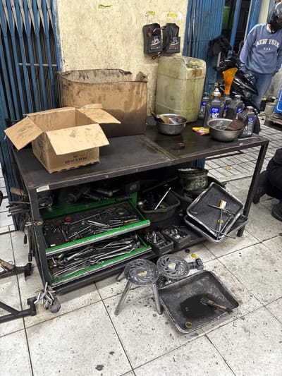 This image captures a cluttered motorcycle repair workshop in Binong, Indonesia, likely in the daytime given the lighting. A person wearing a blue sweatshirt with "NEW YORK U.S.A." printed on it and a helmet is partially visible, sitting on a motorcycle. Various tools and automotive supplies are strewn across a metal cart and the floor, suggesting an ongoing or recently completed repair.

Prominently featured are several containers of oil, including a large translucent jerrycan and smaller black bottles with "X-TEN" branding. A yellow funnel is placed near the motorcycle's engine area. The cart itself is filled with an assortment of tools, including wrenches, pliers, and what appear to be socket sets, housed in green organizers. Several metal trays, some containing used oil and a brush, are also on the cart. Two disc brake rotors are visible on the floor.

In the background, a blue accordion-style gate is closed, and a damaged cardboard box is positioned to the left. The floor is tiled and appears dirty, consistent with a working garage environment. Some faint text is visible on the cardboard box, reading "Isi 120 ml X 24 btl." The overall impression is of a busy, functional repair space with a focus on motorcycle maintenance.